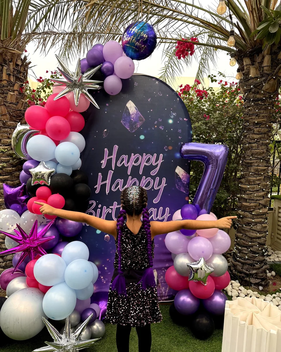 Birthday girl with her arms open in front of a purple K-pop seventh birthday backdrop with balloons