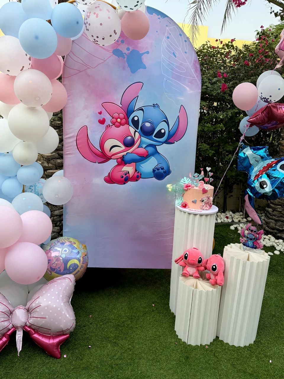 Pastel character themed birthday backdrop with a balloon garland, cake and plush toys