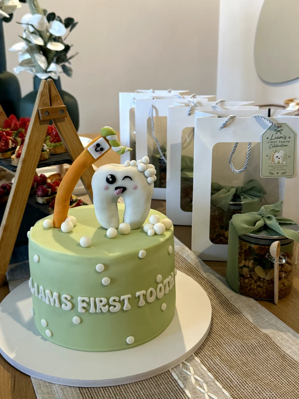 Personalised First Tooth cake with a smiling tooth topper, beside wrapped favour bags