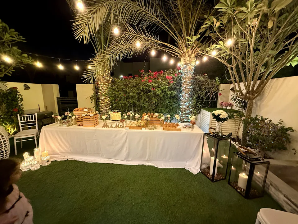 Garden setup for an intimate civil celebration with a long white dessert table under string lights and palm trees