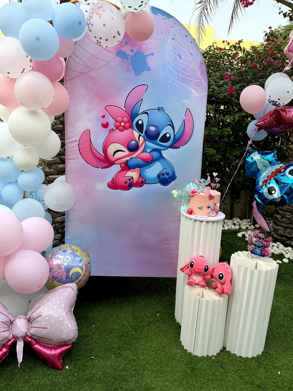 Stitch and Angel birthday backdrop with pastel balloons, a small cake and pink plinths in a garden