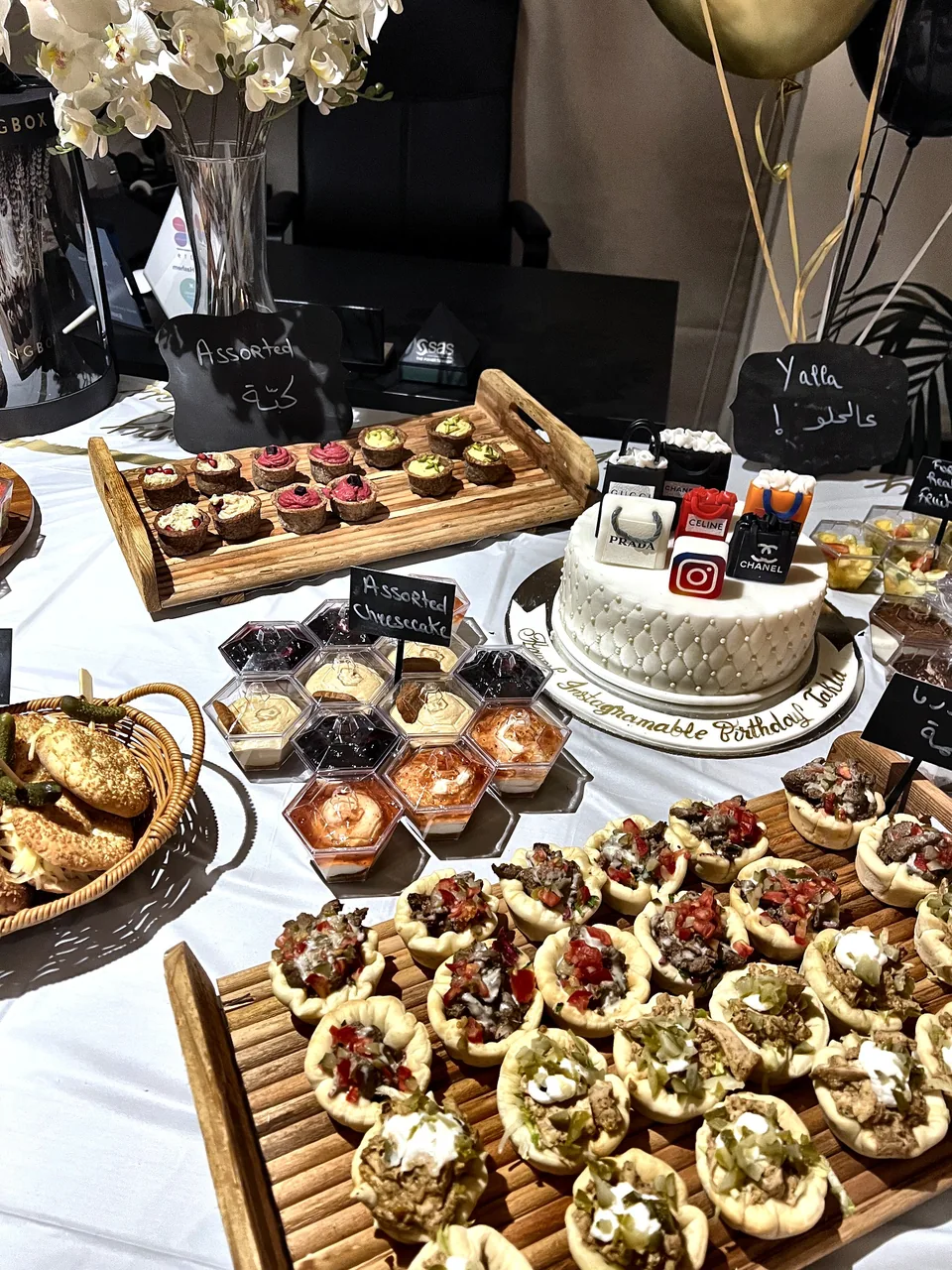 Birthday food table with a cake topped with miniature designer bags, tarts and labelled savoury trays