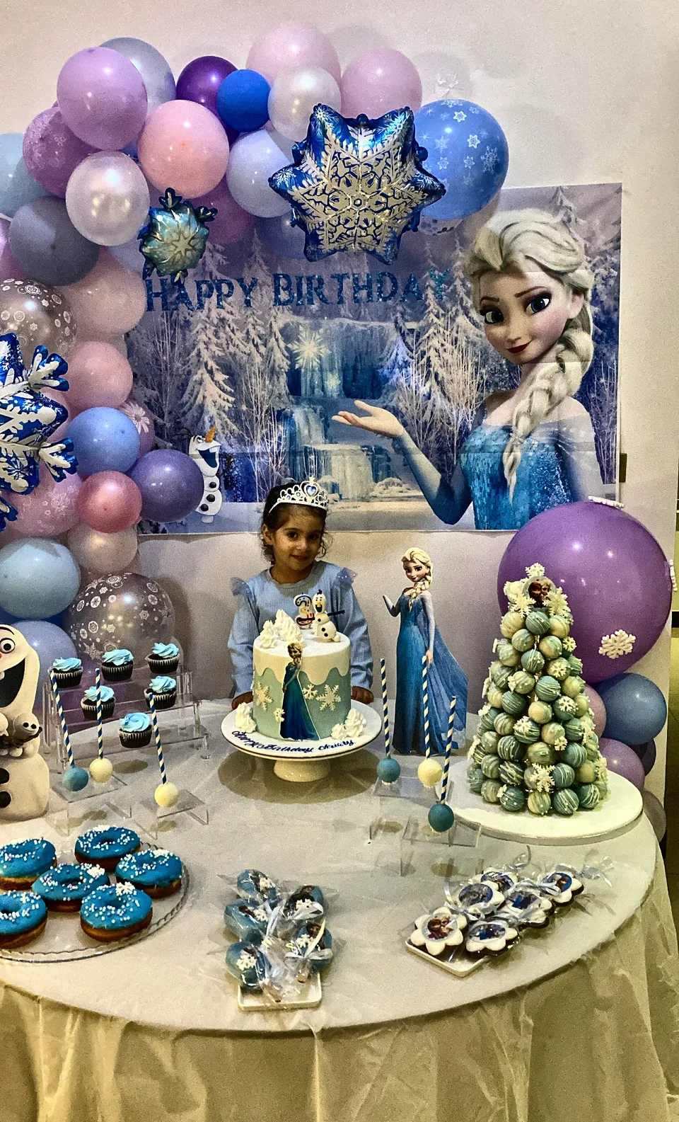 Frozen birthday table with a snowflake balloon arch, an Elsa backdrop, a themed cake and blue treats