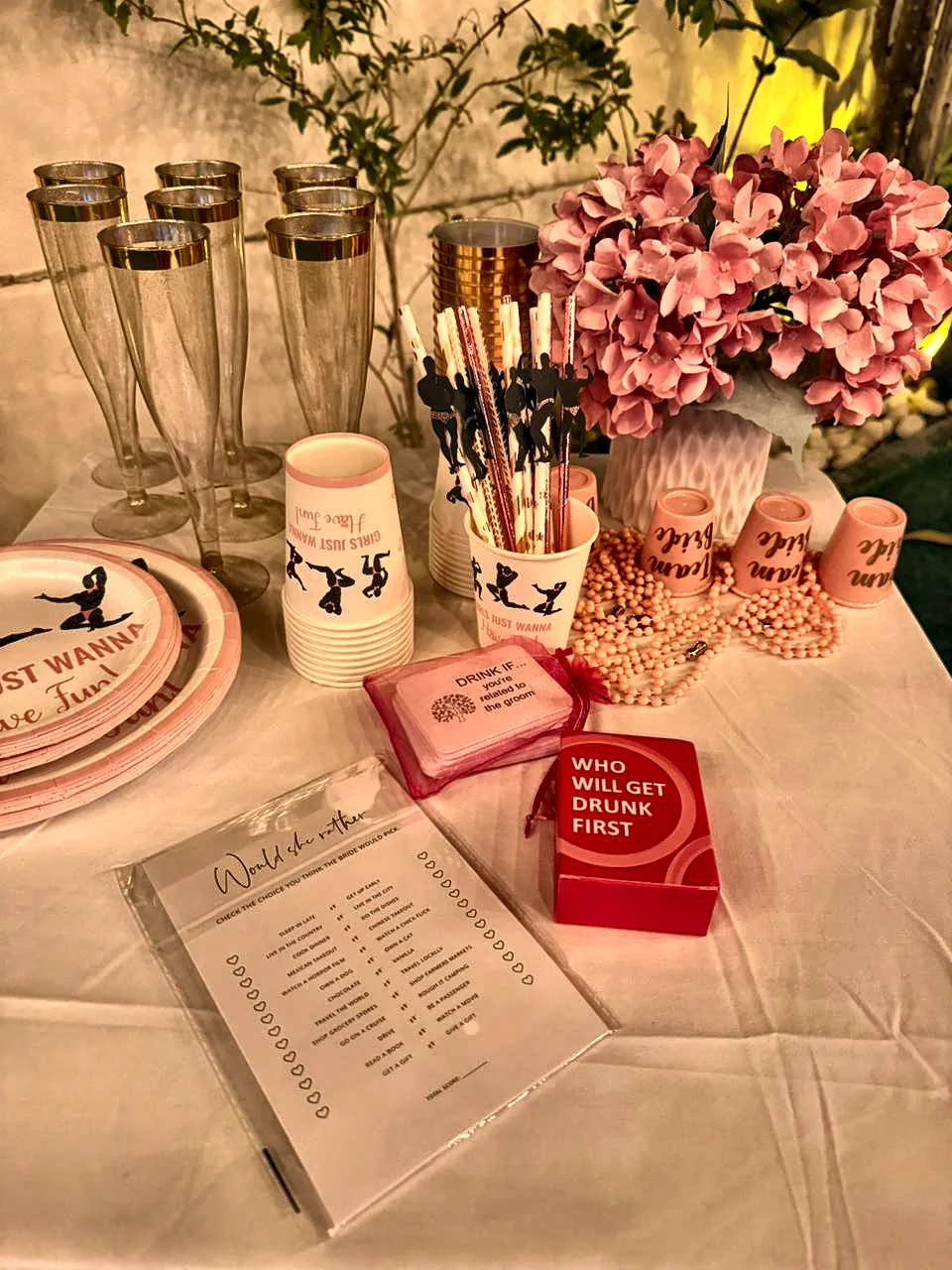 Bachelorette games table with a printed quiz, Who Will Get Drunk First cards, pink flowers and gold-rimmed glasses
